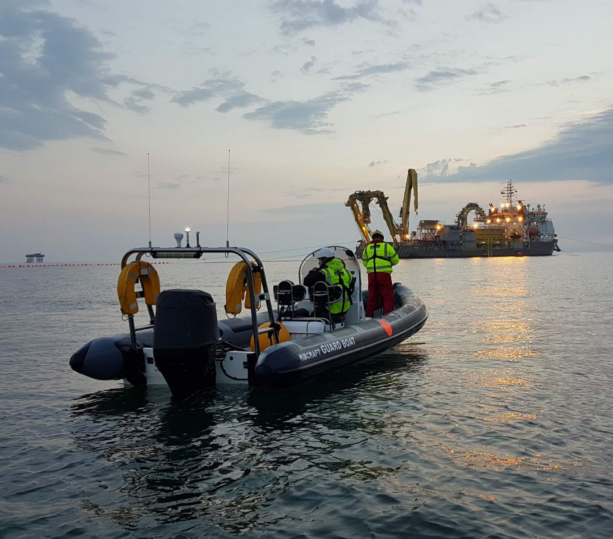 Commercial RIB guard boat operating near offshore construction vessel at dusk, providing safety cover and exclusion zone patrol during marine works in UK waters.